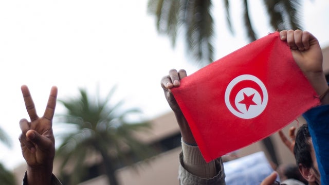 Hands hold up a small Tunisian flag with a peace hand sign to the left