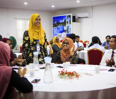 A woman stands and smiles at a table while others clap for her in celebration