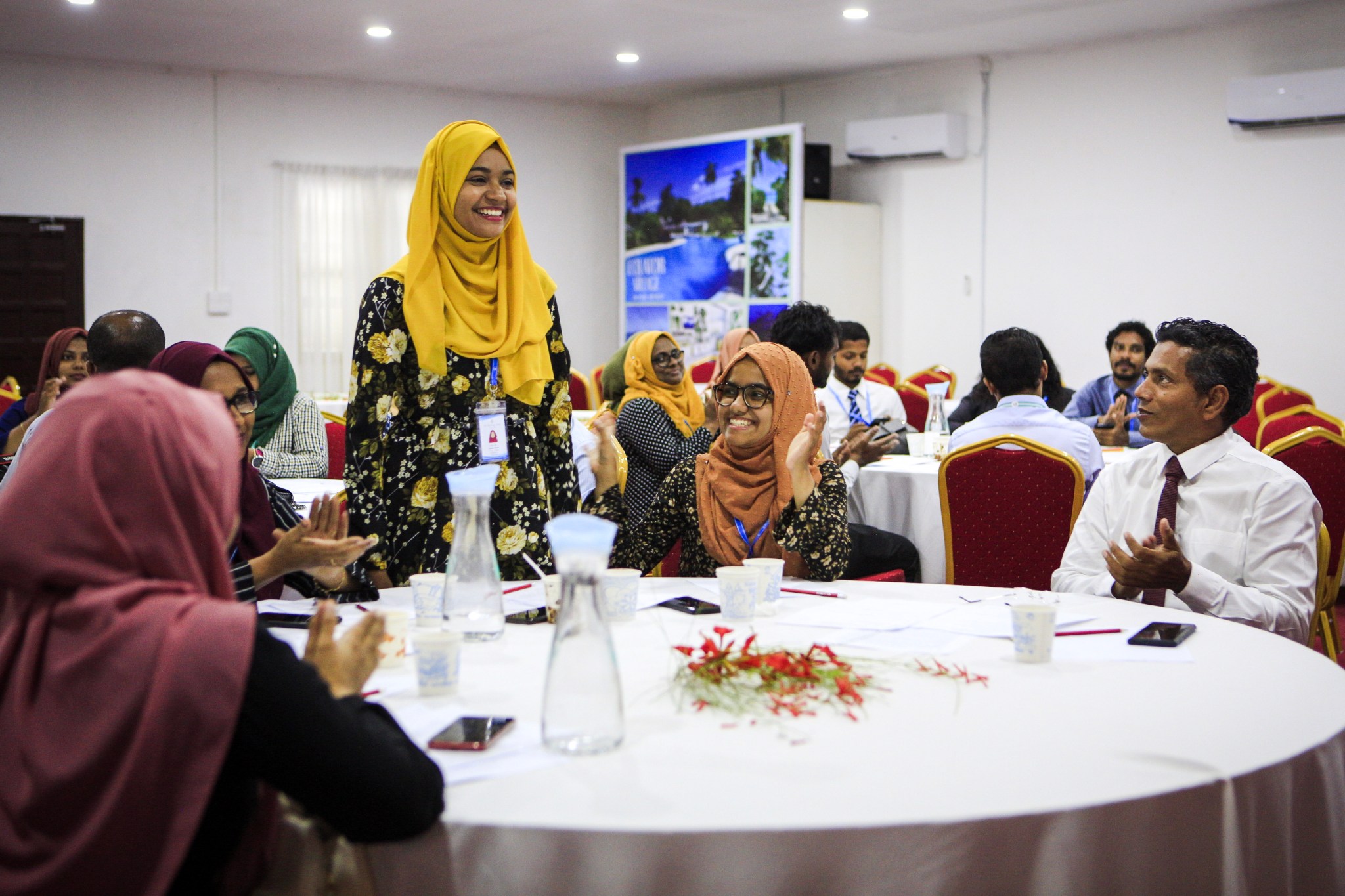 A woman stands and smiles at a table while others clap for her in celebration