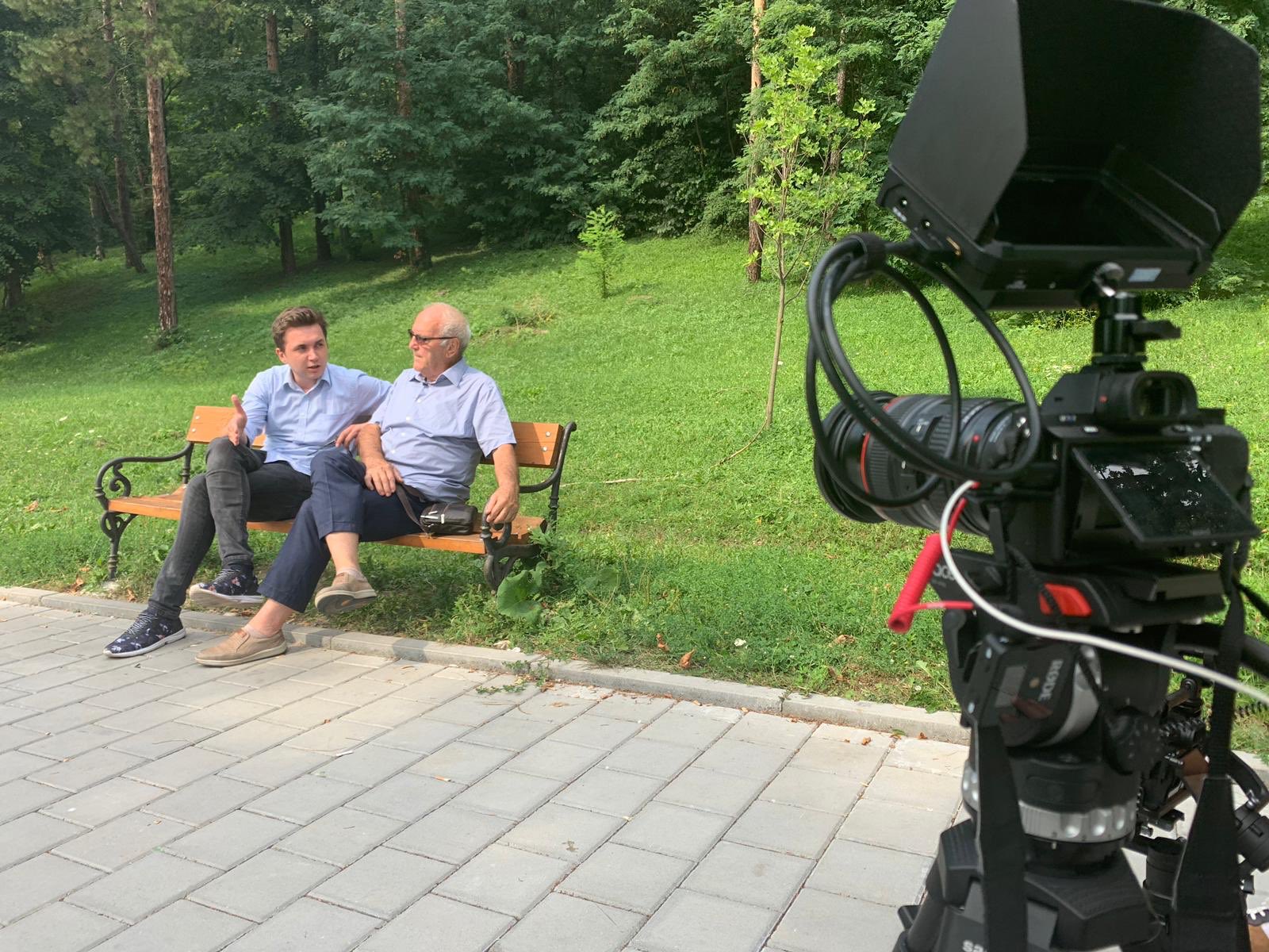 Two men sit on a bench with a camera pointed at them to talk about independent media efforts