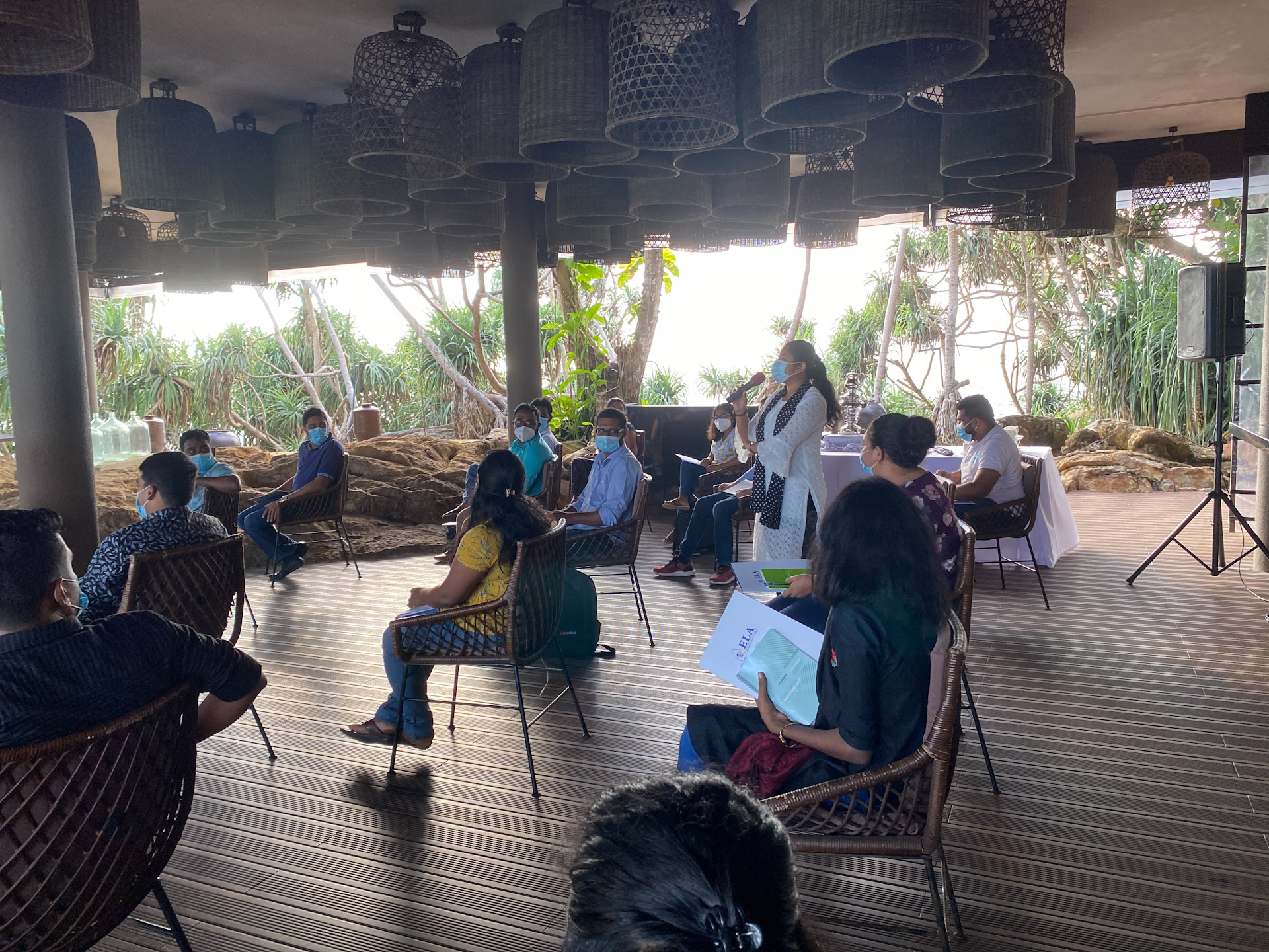 A group sits outside while listening to a lady speak into a microphone in Sir Lanka